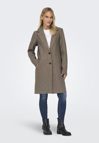 Long manteau en laine brun avec un col cranté, deux boutons noirs et des poches latérales, associé à un jean bleu et des bottines noires.