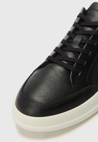 Black leather sneaker with textured upper, round toe, and flat black laces. Features a white rubber sole and subtle stitching details.
