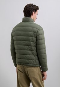 Chaqueta de plumas verde con acolchado horizontal, cuello alto y una textura suave, combinada con pantalones caqui que tienen bolsillos traseros.