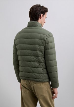 Chaqueta de plumas verde con acolchado horizontal, cuello alto y una textura suave, combinada con pantalones caqui que tienen bolsillos traseros.