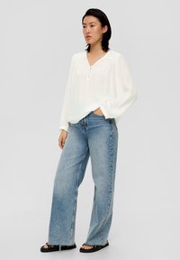 Blouse blanche à manches longues avec un décolleté en V et des détails de boutons, associée à un pantalon en jean large bleu clair et des sandales noires, sur un fond neutre.