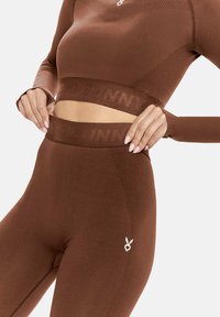Barna sportos szett, testhezálló, hosszú ujjú felsővel és magas derekú leggings-szel. Sima textúra, logó részletekkel és hálós betétekkel.