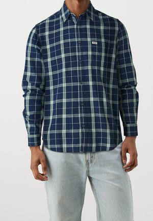 Homme portant une chemise à carreaux bleu marine et vert à boutons avec le logo Wrangler sur la poche poitrine et un jean bleu clair, debout devant un fond blanc.