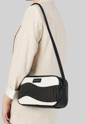 Femme portant un blazer beige, tenant un sac à bandoulière noir et blanc avec un motif texturé et une longue sangle sur fond gris.