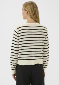 Femme aux longs cheveux blonds portant un pull crème avec des rayures horizontales noires et un pantalon noir, vue de dos.