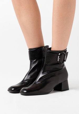 Botines negros de tobillo hechos de piel brillante, con un cierre de cremallera, un puño con una hebilla redonda y un tacón bajo tipo bloque.