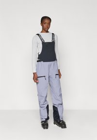 Smučarske kombinezoni z črnim zgornjim delom in svetlo sivo spodnjo polovico, narejeni iz teksturiranega materiala. Vključujejo nastavljive naramnice in žepe z zadrgo.