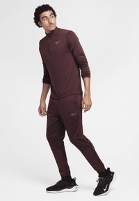 Brązowa koszulka sportowa Nike z półzamkiem, zestawiona z dopasowanymi brązowymi spodniami. Czarne sneakersy Nike z białą podeszwą. Gładka, dopasowana faktura materiału.