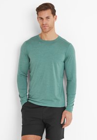 Man standing against white wall wearing a teal long-sleeve shirt and black shorts with one hand in pocket.