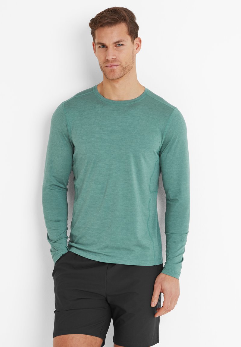 Man standing against white wall wearing a teal long-sleeve shirt and black shorts with one hand in pocket.