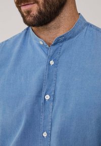 Camisa de chambray azul claro con cuello tipo banda y bolsillos aplicados, con botones blancos y una tela suave con ligera textura.