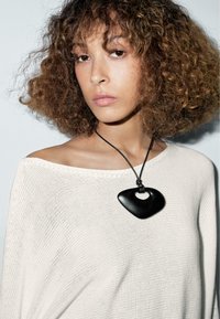 Black pendant necklace with a unique heart-shaped design, hung on a black cord, worn over a light knitted sweater.