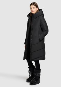 Frau in einem langen schwarzen Daunenmantel mit hohem Kragen und schwarzen Schneestiefeln, die vor einem einfarbigen hellgrauen Hintergrund steht.