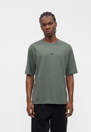 Ung mand med flettet hår står med fronten mod kameraet iført en løs grøn t-shirt og sorte bukser mod en ensfarvet lys baggrund.
