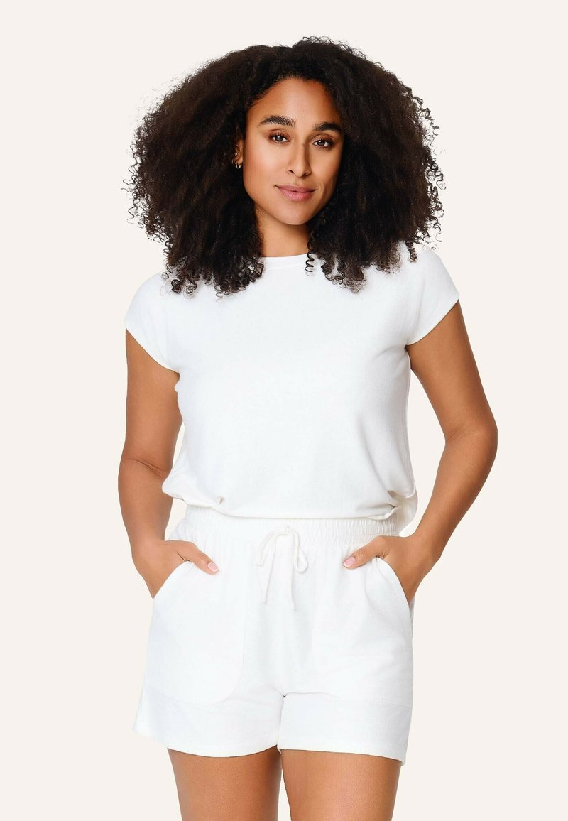 Femme aux cheveux bouclés et foncés portant un haut blanc à manches courtes et un short à cordon, debout les mains dans les poches devant un fond clair.