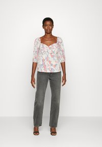 Blusa com padrão floral, apresentando um decote quadrado, mangas curtas com volume e um tecido leve. Combinada com jeans cinzentos de cintura alta e saltos.