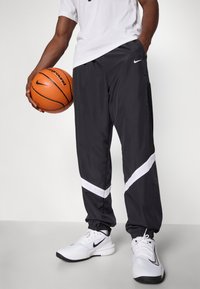 Calças de desporto pretas com detalhes brancos, feitas de material leve. Apresentam punhos elásticos e bolsos laterais em malha. O jogador segura uma bola de basquete.
