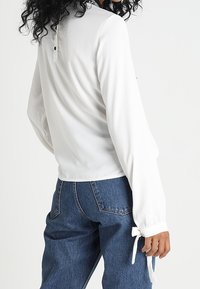 Femme aux cheveux bouclés portant un chemisier blanc à manches longues avec fermeture boutonnée dans le dos et poignets noués, associé à un jean bleu taille haute.