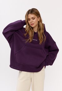 Felpa oversized viola realizzata in tessuto morbido, con tasca frontale, cappuccio con coulisse e cuciture decorative sui bordi.
