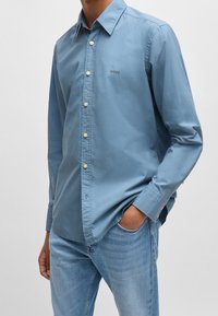 Camisa de botón de color azul claro en algodón, con cuello clásico, mangas largas y un pequeño detalle de logo en el pecho.