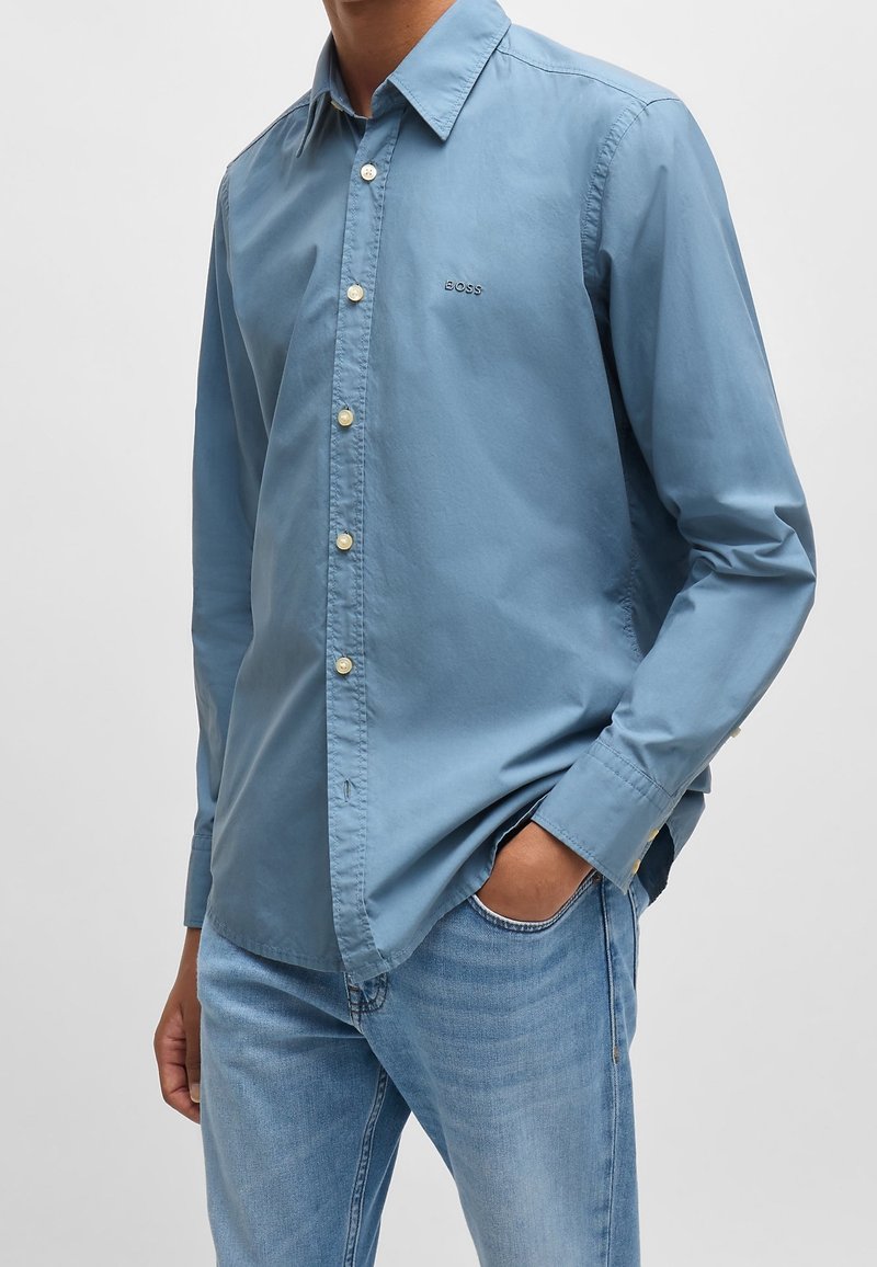 Camisa de botón de color azul claro en algodón, con cuello clásico, mangas largas y un pequeño detalle de logo en el pecho.