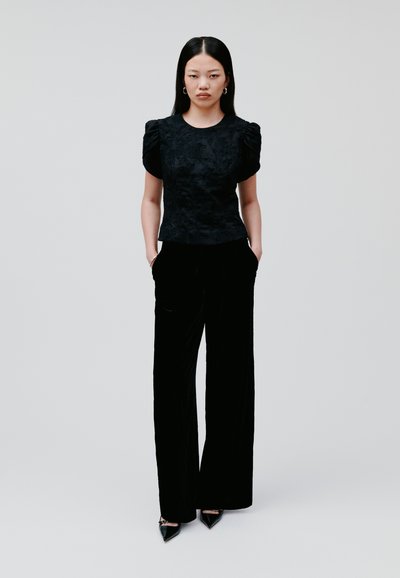 Schwarze strukturierte Bluse mit kurzen Puffärmeln, kombiniert mit hochgeschnittenen schwarzen Samt-Hosen. Das Model steht mit den Händen in den Taschen.