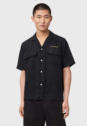 Młody mężczyzna ubrany w czarną, krótki rękaw, koszulę zapinaną na guziki z dwoma kieszeniami na piersi i haftem "ALLSAINTS" w kolorze złotym.