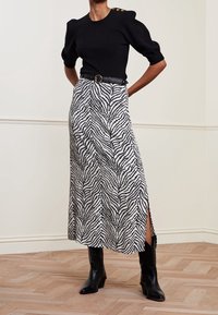 Zwart gebreide top met korte pofmouwen en gouden knoopaccenten, gecombineerd met een witte rok met zwart golvend patroon en zijopeningen.