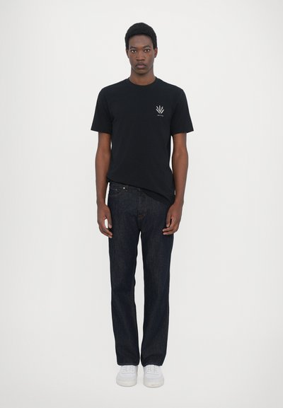 T-shirt noir à manches courtes avec un design graphique sur la poitrine, associé à un jean en denim bleu foncé et des baskets blanches.