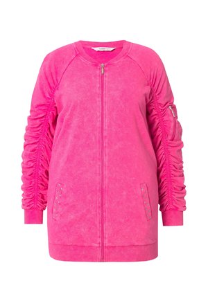 Veste zippée rose avec des poignets côtelés, présentant des détails froncés sur les côtés et une poche zippée. Fabriquée en tissu doux et texturé.
