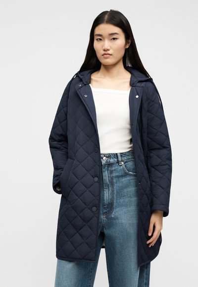 Νεαρή γυναίκα με μακριά σκούρα μαλλιά, φοράει ναυτικό μπλε καπιτονέ παλτό με κουκούλα πάνω από λευκό τοπ και ψηλόμεσα μπλε τζιν, στέκεται μπροστά σε απλό φόντο.