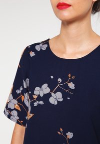 Robe bleu marine avec un motif floral en gris clair et orange, présentant un col rond et des manches courtes. Tissu lisse.