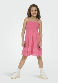Růžové bavlněné šaty se smyčkovým živůtkem, sukni ve vrstvách s okenními výřezy a nastavitelnými ramínky. V kombinaci s bílými teniskami a ponožkami do kotníků.