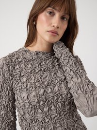 Femme jeune aux cheveux châtain clair portant un haut long à manches avec une texture grise légèrement froncée, posant une main près de son cou contre un fond blanc.