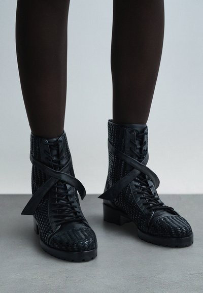 Svarta läderstövletter med snörning, vävd struktur, platt klack och dekorativa remmar runt fotleden, med mönstrade detaljer på tån och sidan.