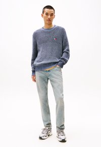 Blauwe gebreide trui met een ronde hals en ribgebreide manchetten, gecombineerd met lichte denim jeans. Sneakers in een wit en grijs kleurenschema.