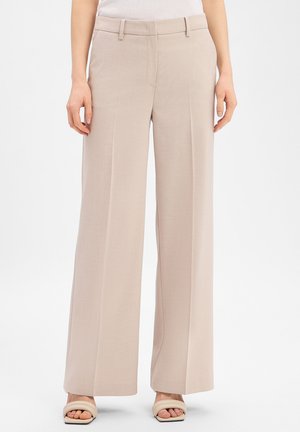 Woman wearing beige wide-leg trousers with front creases, belt loops, and beige open-toe sandals, standing against a plain background.