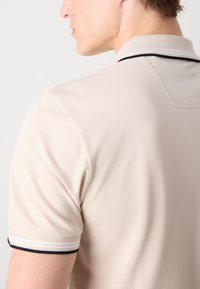 Beige pólóing fekete és fehér csíkozott bordás gallérral. Az anyag sima tapintású és testhezálló szabású.