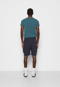 Tílko s krátkým rukávem v barvě modro-zelené, kombinované s tmavě modrými cargo šortkami, které mají boční kapsy a volnější střih. Bílé sportovní ponožky a nazouvací boty.