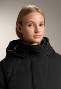 Fekete pufidzseki magas nyakkal és kapucnival. Az anyaga sima, steppelt textúrájú. Elöl cipzárral rendelkezik, és karcsúsított fazonú.