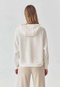 Felpa color crema con una texture morbida, dotata di cappuccio con cordino, vestibilità comoda e polsini a coste. Indossata con pantaloni beige chiaro.