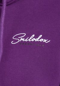 Lilla sweatshirt med blødt, tekstureret stof. Forsiden viser mærkenavnet "Smilodox" i hvid kursiv med "ESSENTIALS" i grøn.