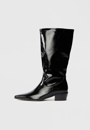 Juodas lakinės odos iki kelių botas su smailiu padu, šiek tiek siaurėjančiu kulnu ir lygia, blizgia apdaila.
