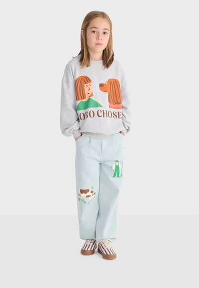 Kind steht mit Händen in den Taschen, trägt einen grauen Pullover mit Hunde- und Mädchenillustration sowie hellblaue Hosen mit landwirtschaftlichen Motiven.