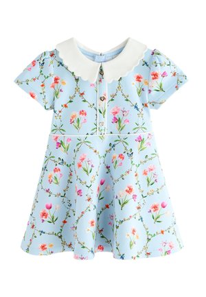 Fată tânără zâmbitoare cu păr creț, purtând o rochie florală albastru deschis cu guleraș alb și pantofi albi cu funde roz, stând pe podea gri.