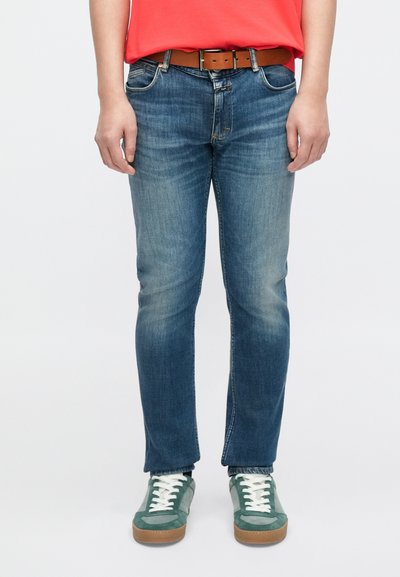 Jean bleu slim avec légère décoloration sur les cuisses, associé à une ceinture marron et des baskets vertes à lacets blancs.