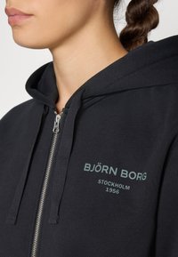 Sweatshirt preto com fecho em zíper e acabamento texturizado. Apresenta capuz, zíper prateado e impressão do logotipo verde claro com a inscrição "BJÖRN BORG STOCKHOLM 1956."