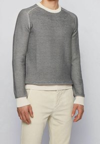 Pull gris en tricot avec un motif texturé, col et poignets côtelés de couleur crème, coupe décontractée, porté avec un pantalon crème.