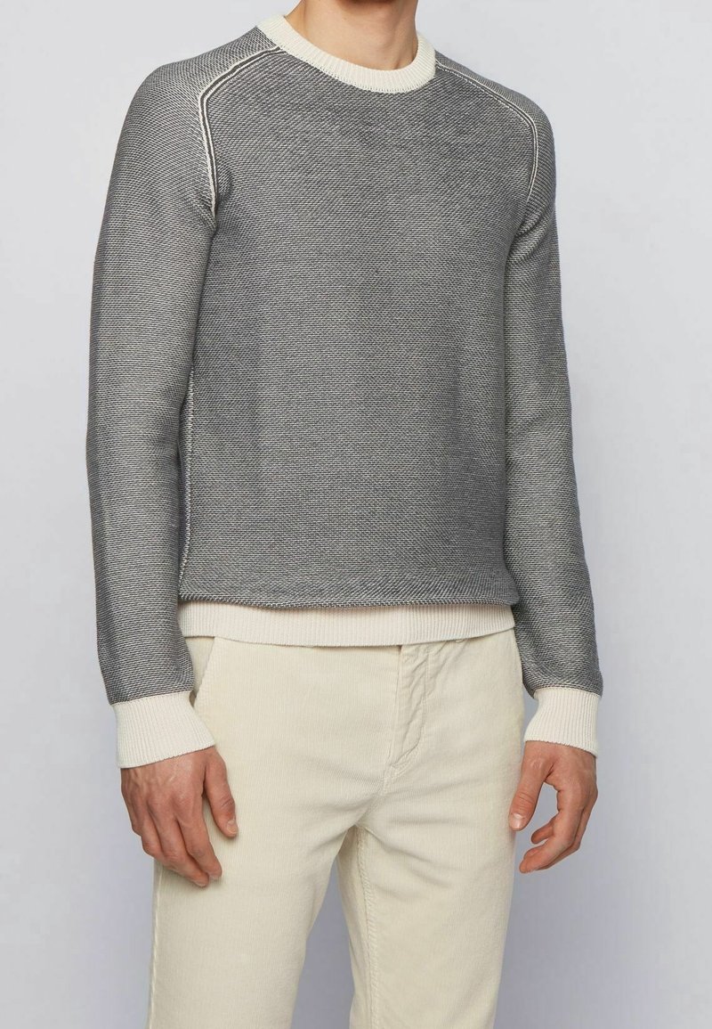Pull gris en tricot avec un motif texturé, col et poignets côtelés de couleur crème, coupe décontractée, porté avec un pantalon crème.