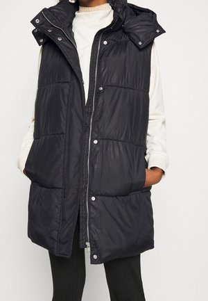 Femme portant un gilet matelassé noir sans manches par-dessus un haut blanc à manches longues, les mains dans les poches du gilet, sur un fond uni.